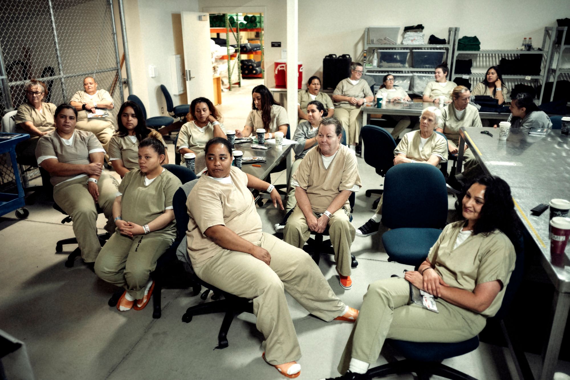 A group of women in tan prison uniforms sitting in a room, watching a television show.