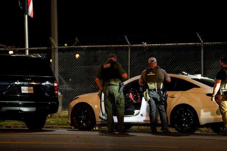 A photo shows officers with vests that read "trooper" and "U.S. Marshal" standing in front of a white car with the door open at night. 