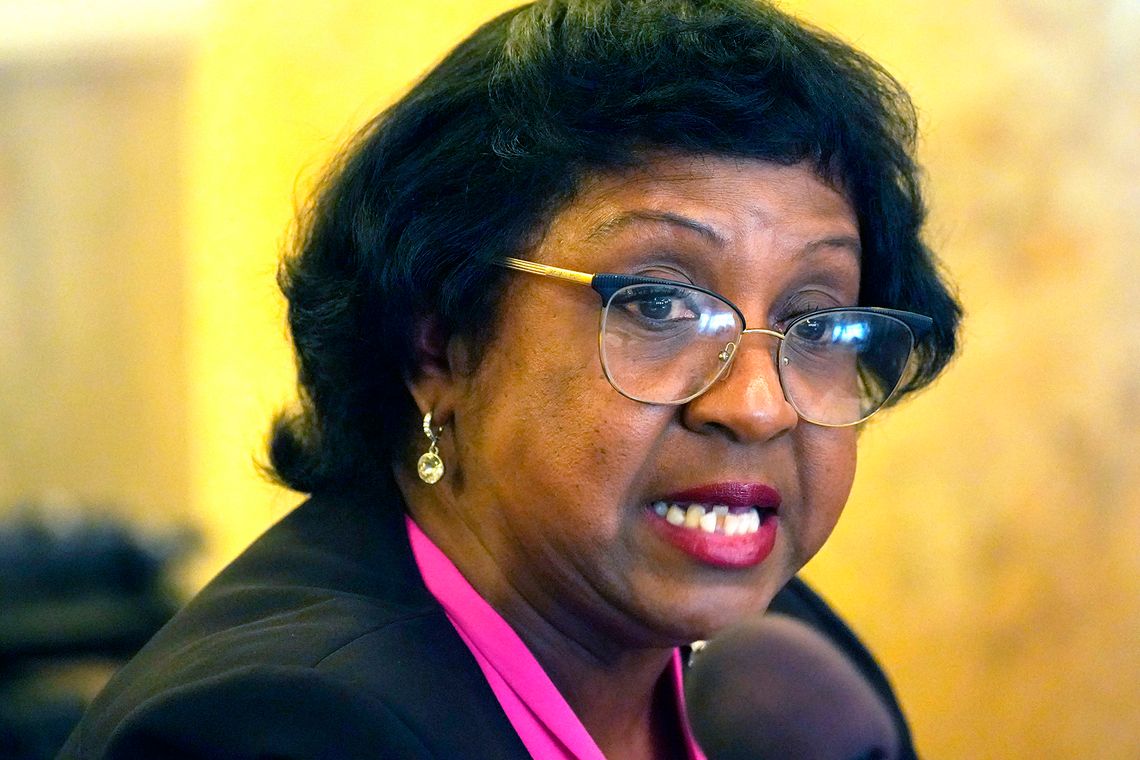 A Black female public defender, wearing glasses, pink blouse and black blazer, speaks into a courtroom microphone.
