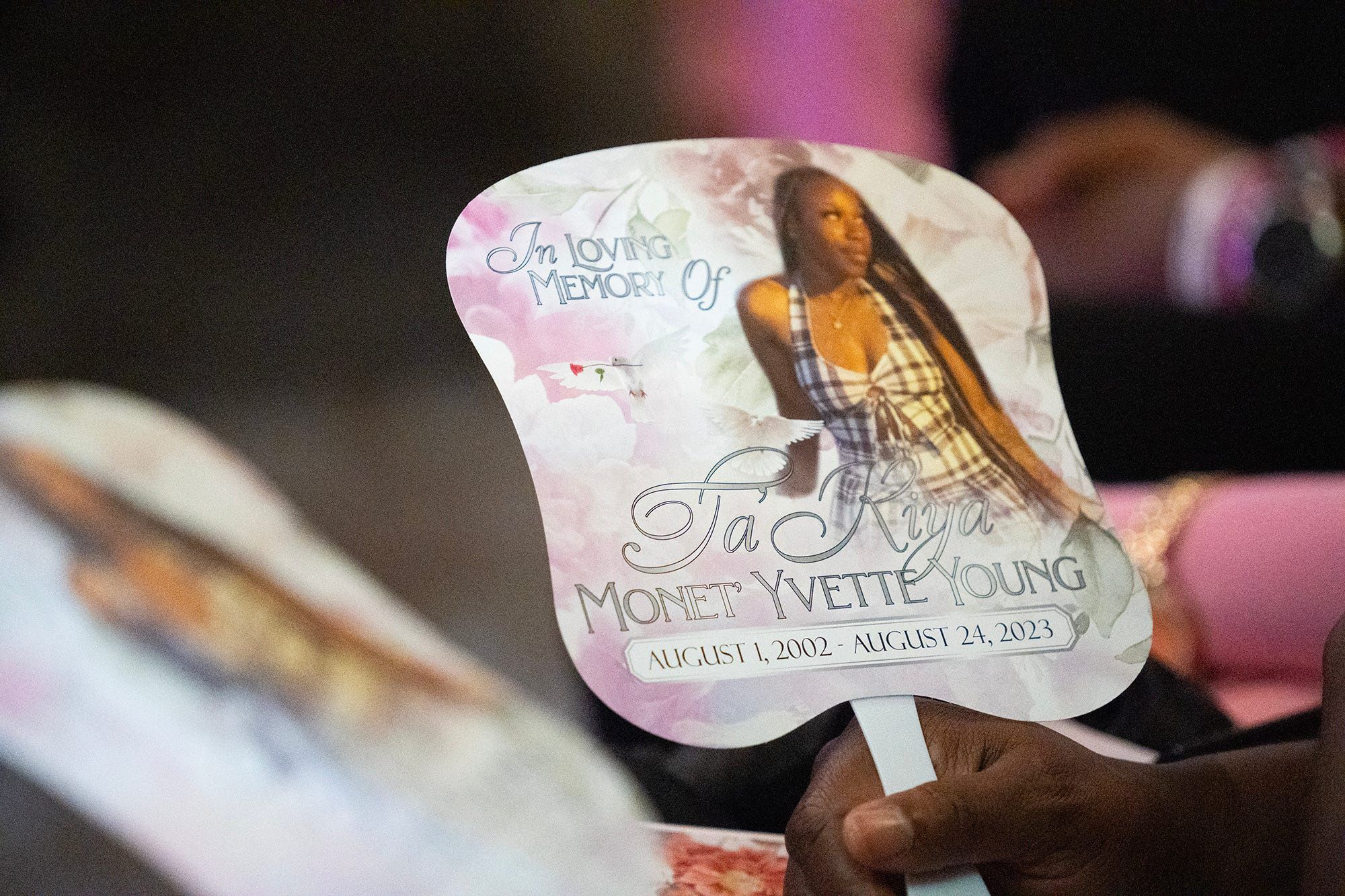 The hand of a Black person holds a fan of an image of a Black woman wearing a black and white plaid dress. The fan reads: "In loving memory of Ta'Kiya Monet Yvette Young. August 1, 2002 - August 24, 2023." 