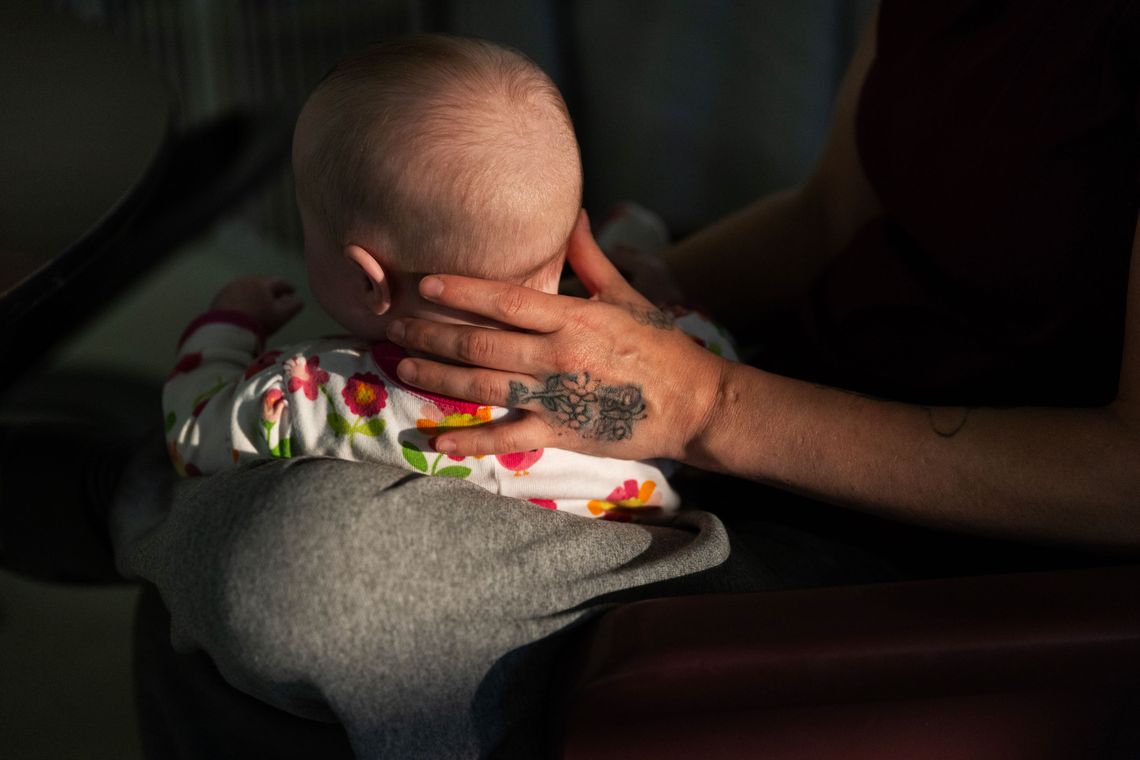 A close-up photo shows a White woman’s hand with a flower tattoo supporting a baby’s head.