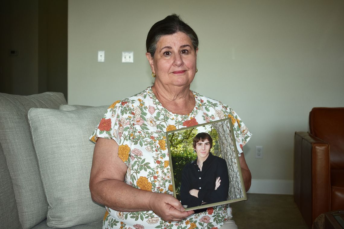 Sue Phillips, who has light-toned skin and wears a floral shirt, faces the camera. She holds a picture of her deceased son, Matthew. 