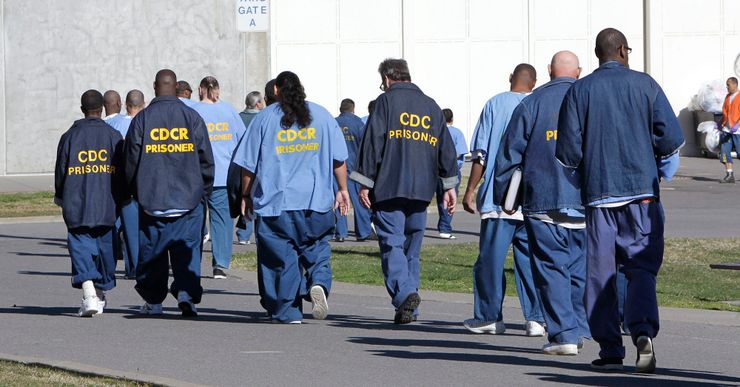 A group of people, wearing blue prison uniforms with the words "CDCR prisoner" on their backs, walk through a prison yard. 