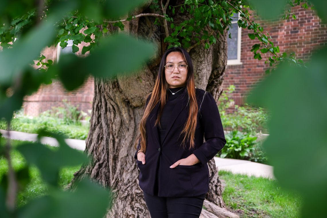 A woman with light brown hair and glasses in a black suit jacket stands in front of a tree.