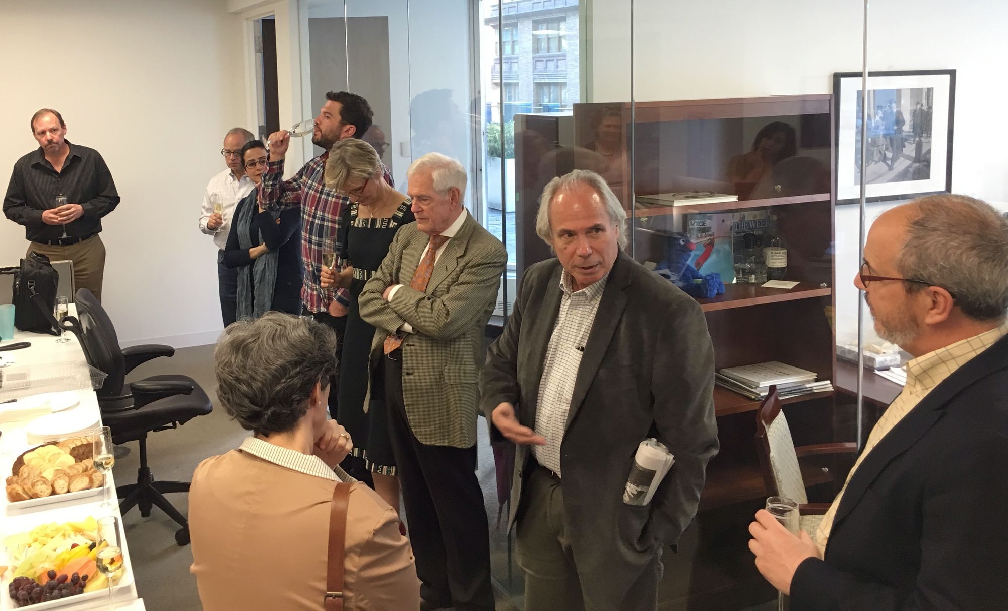 Tom Robbins, a man with light-toned skin and gray hair, wears a suit jacket and stands in a newsroom with eight other people. Behind them are offices visible through glass walls.  