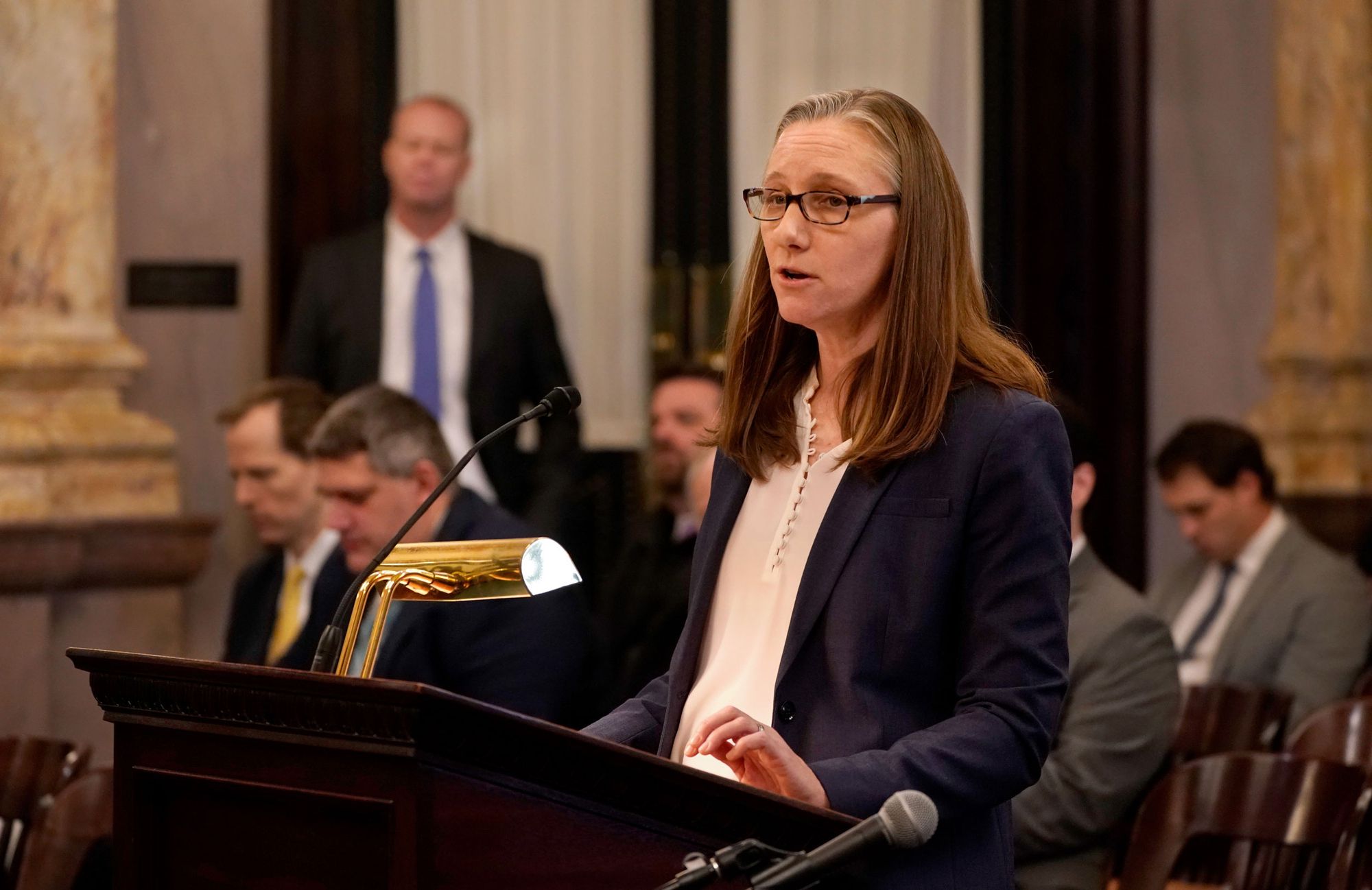 A White woman with glasses speaks at a podium. 