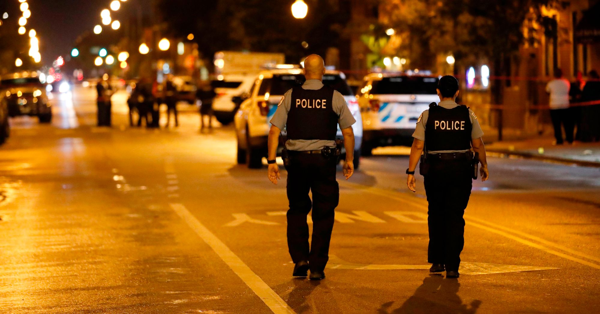 Two police officers walk in the middle of a street at night toward the scene of a shooting. 