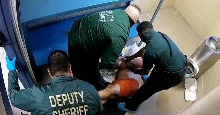 A man in orange prison shorts is being restrained by three officers in a prison cell. 