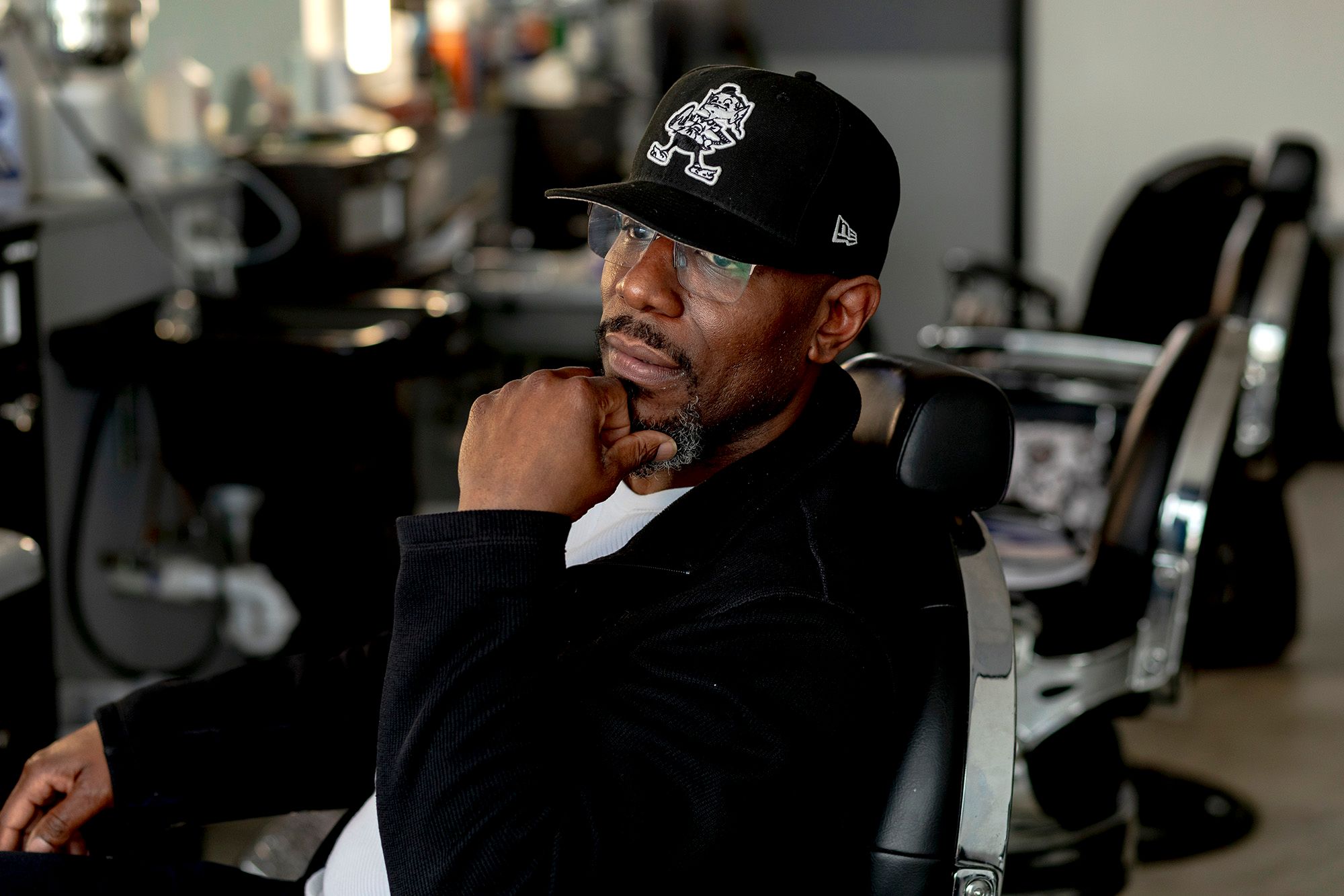 A Black man, in a black jacket and white shirt, poses for a portrait while sitting on a barber chair. 