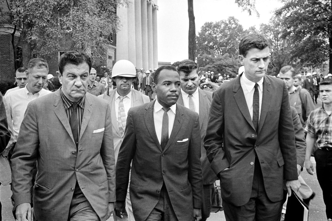A black and white photo of a Black man in a suit walking through a college campus while surrounded by a group of White men in suits. One of the White men is wearing a helmet. 