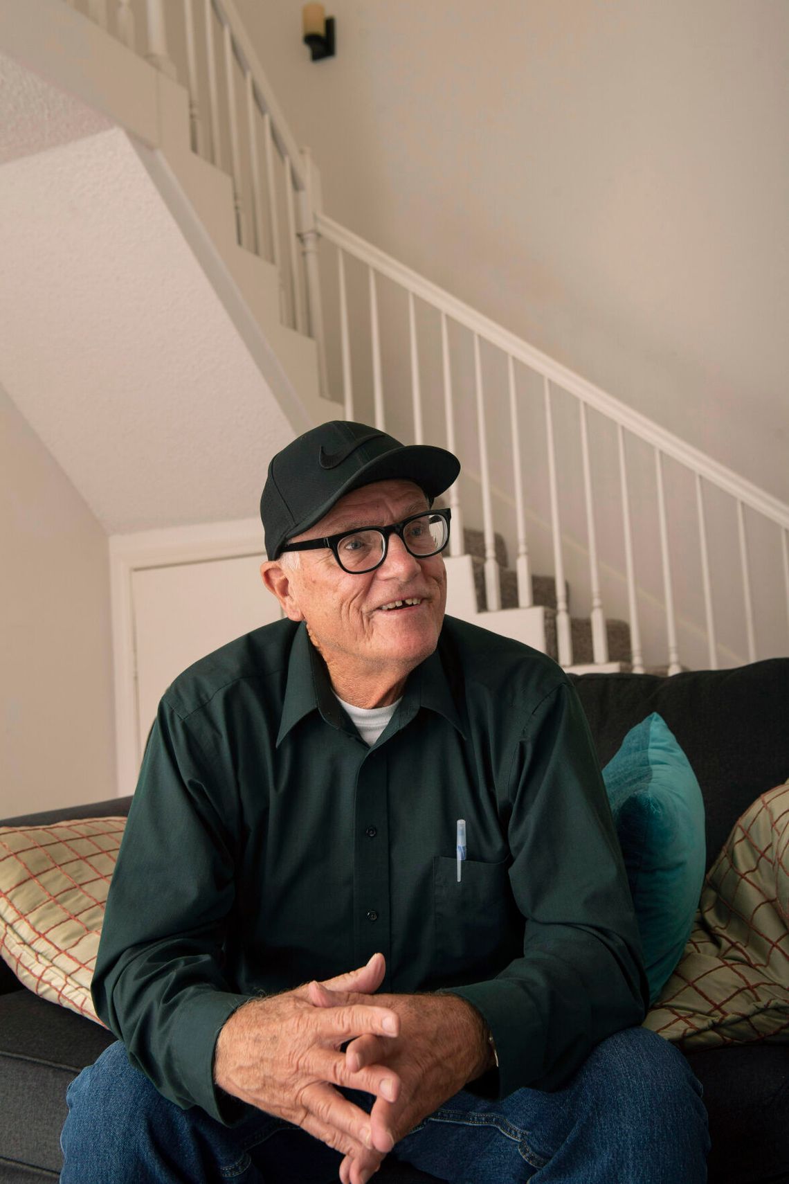 Kenneth Womack, an elderly man, wears a long-sleeved green shirt and wears dark-rimmed glasses and a baseball hat. 