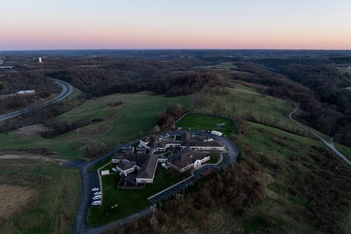 An aerial photo shows a cluster of buildings with grey roofs surrounded by green hills, and a road to the left. 