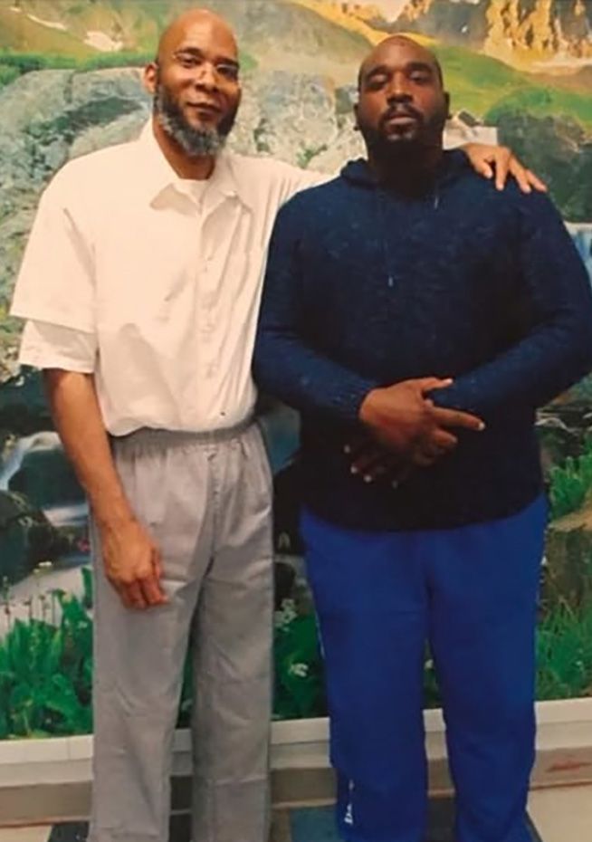 A Black man in a blue sweatshirt and pants stands next to his father for a picture, who is wearing a white prison uniform. 