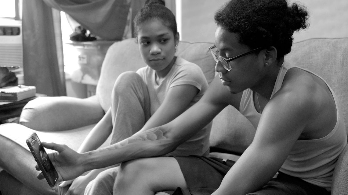 A teenage boy looks at his phone during a FaceTime video call. His younger sister looks at him while sitting next to him on a couch. 