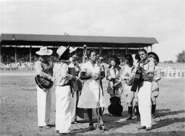 A black-and-white photo of a group of musicians dressed in white, with some wearing cowboy hats, standing in a horseshoe shape, while a Black woman in a white dress sings into a microphone. The group is standing on the rodeo grounds, and in the background are the stands where the audience is seated. 
