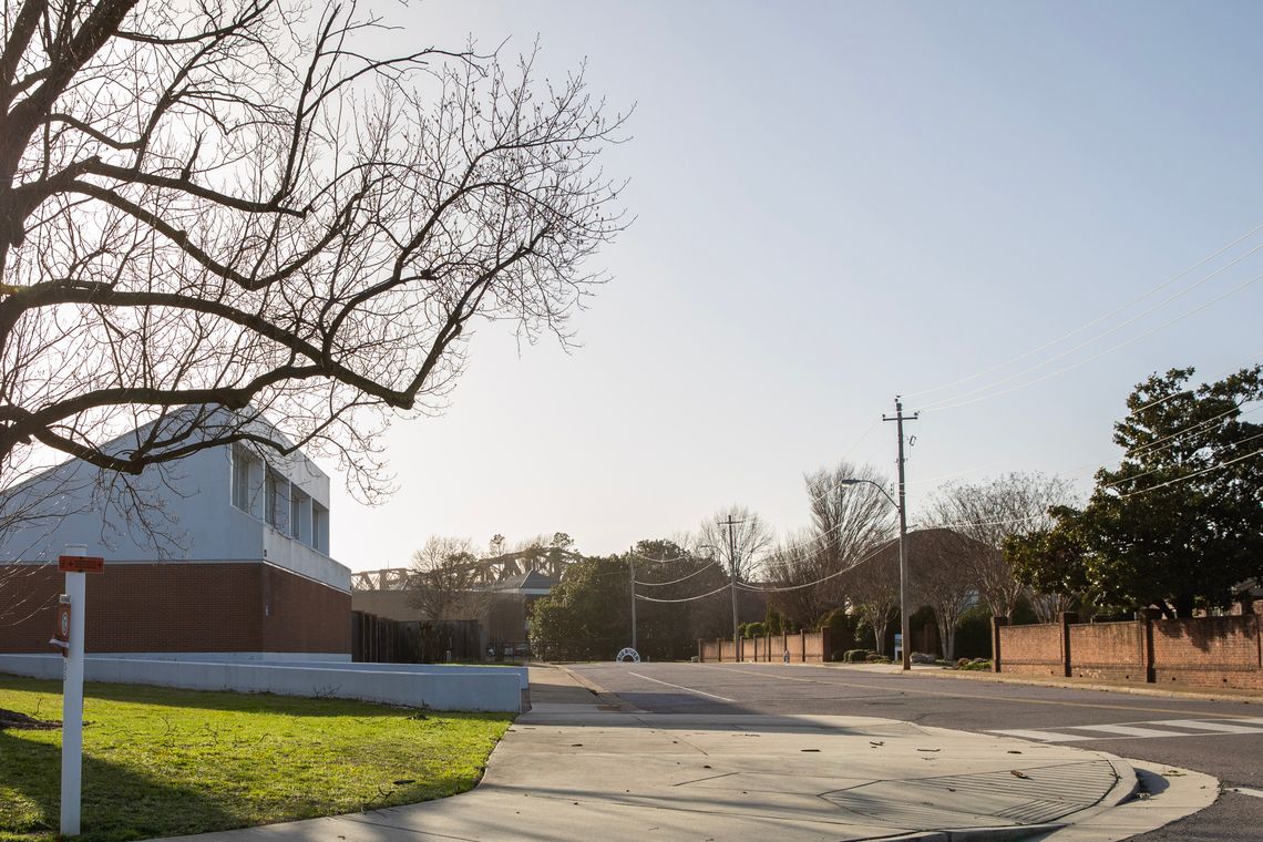 A photo shows a white and brown building on the left side of the frame, next to a sidewalk and street on the left during the daytime. 