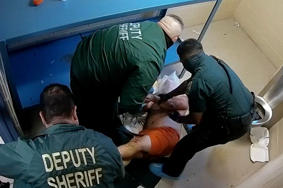 A man in orange prison shorts is being restrained by three officers in a prison cell. 