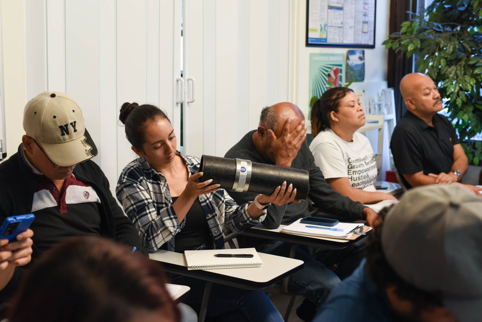 A group of five people representing a mix of genders and ethnicities sit at desks in a classroom facing the same direction. One woman holds a large black pipe. She is examining the pipe, with her head cocked to the side.