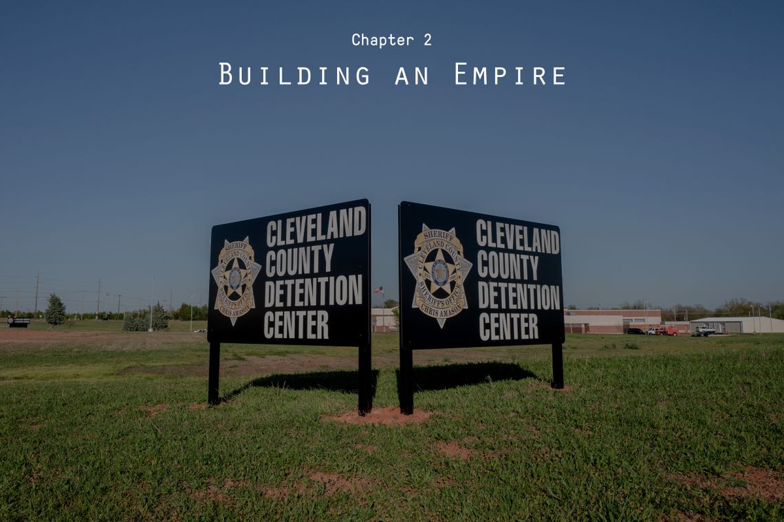 Two dark-colored signs with the words “Cleveland County Detention Center” stand next to each other in a field of grass and beneath a blue sky. The words “Chapter 2: Building an Empire” hover in the sky. 