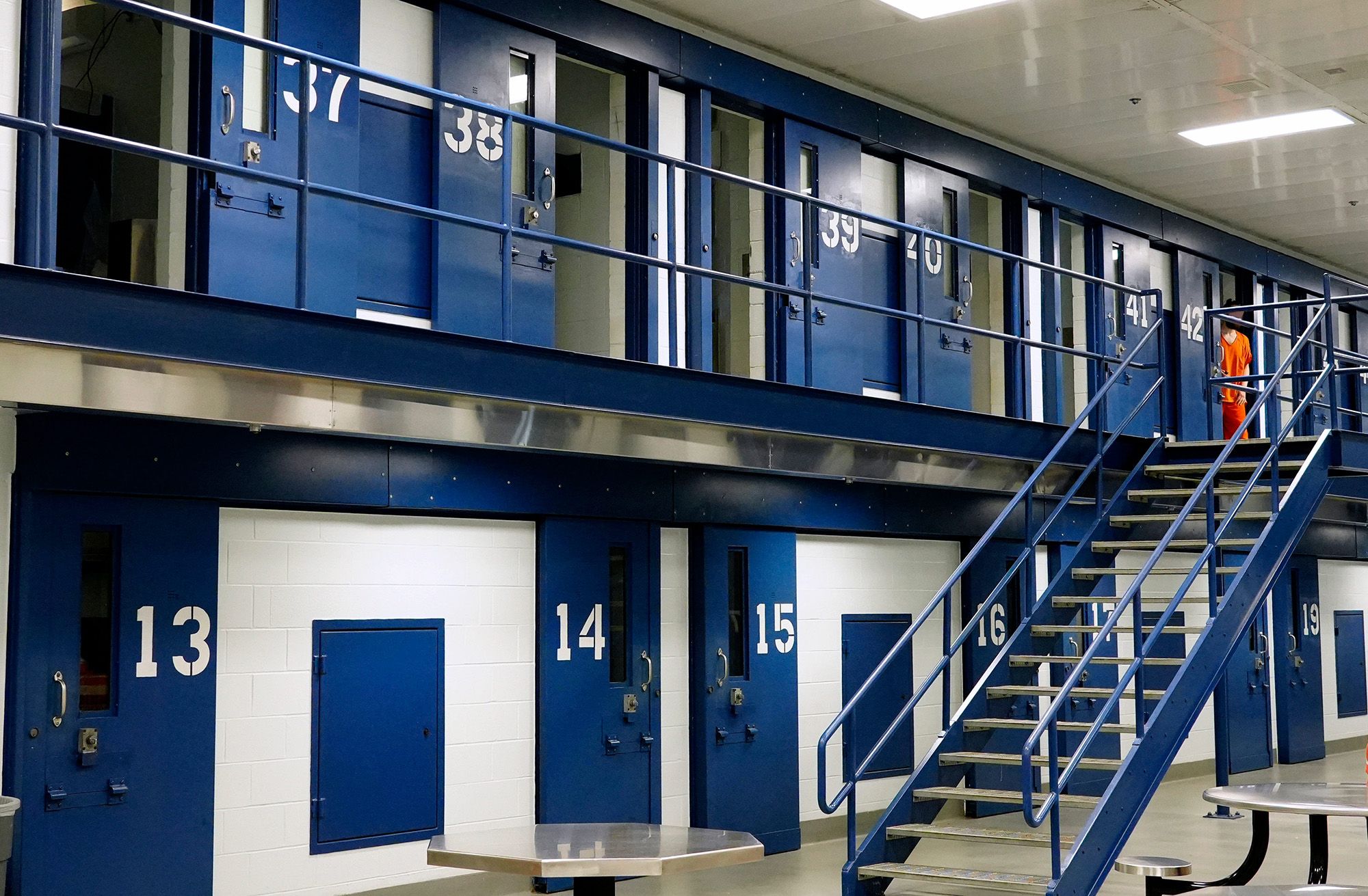 An interior shot of Butler County Jail shows two floors of cells. Each cell door is blue and numbered individually. A person in an orange uniform steps out of their cell. 