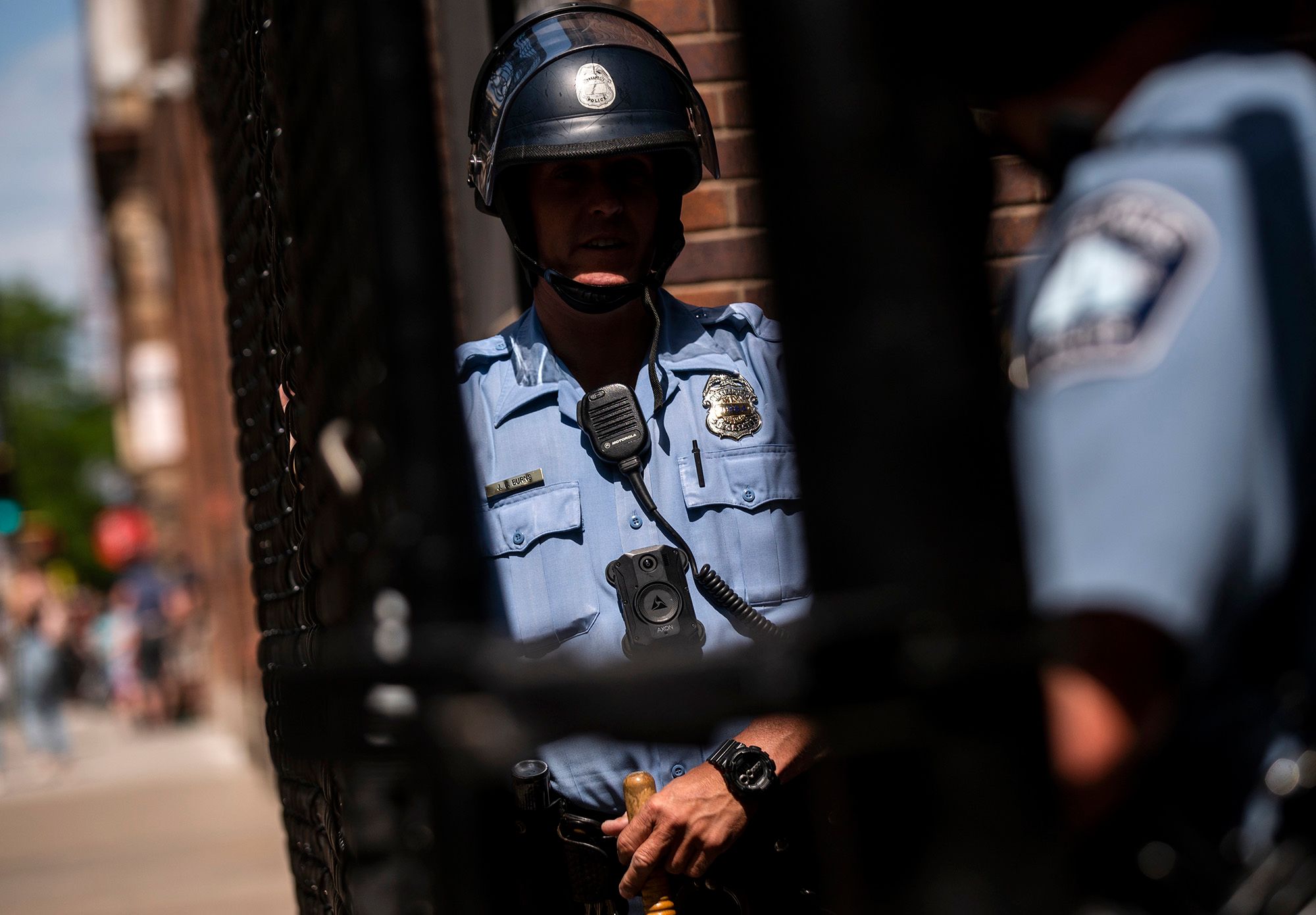 A photograph shows two police officers standing next to a gate on a street. One officer is wearing a helmet and holds a wooden baton. 