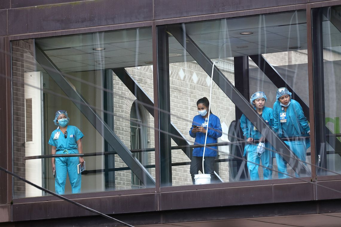 Nurses, wearing blue uniforms, stand in a hospital walkway, looking out a window. 