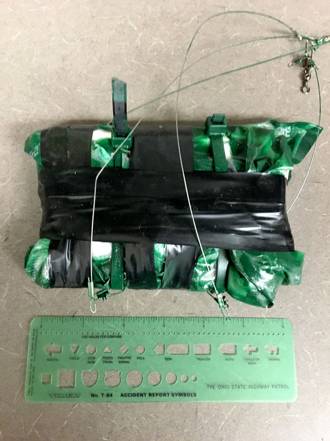 A photo shows a package that was brought by a drone. The package is wrapped in green plastic and black tape. A ruler is situated below the package. 