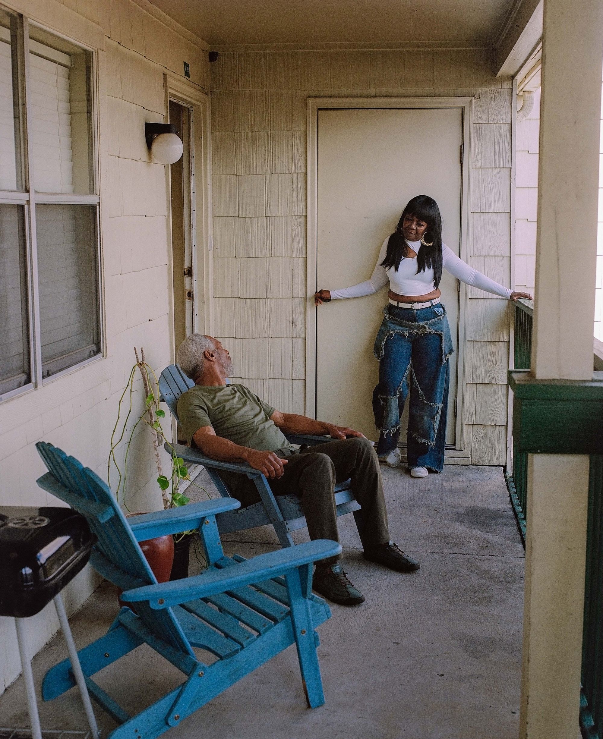 Moses, a Black man wearing an olive green T-shirt and pants, sits in a blue Adirondack chair, while Armour, a Black woman wearing dark patchwork jeans and a white midriff shirt with a chest cutout, leans against a door on the porch of their apartment. 