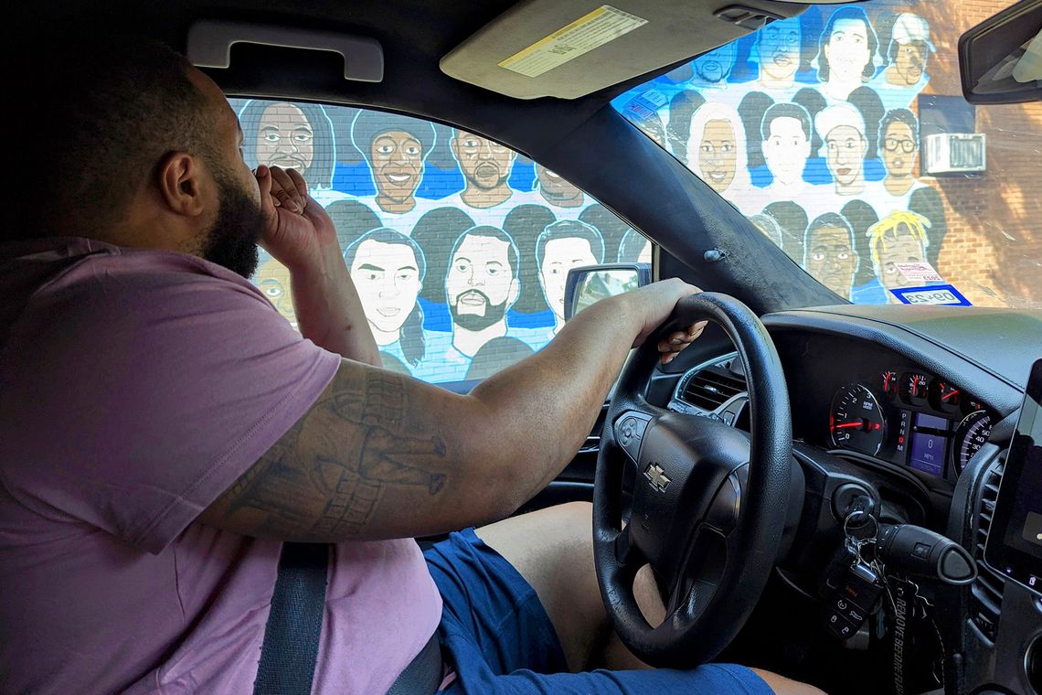 A Black man, wearing a light purple T-shirt and blue shorts, sits in the driver’s seat inside his car. He looks through his window at a mural of multiple portraits. 