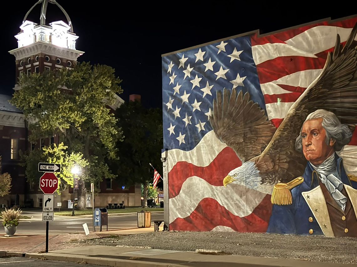 A streetscape of a downtown, with a tall tower standing in the left of the frame. A mural with an American flag, an eagle and George Washington adorns a brick wall and sits on a street corner in the right side of the frame.