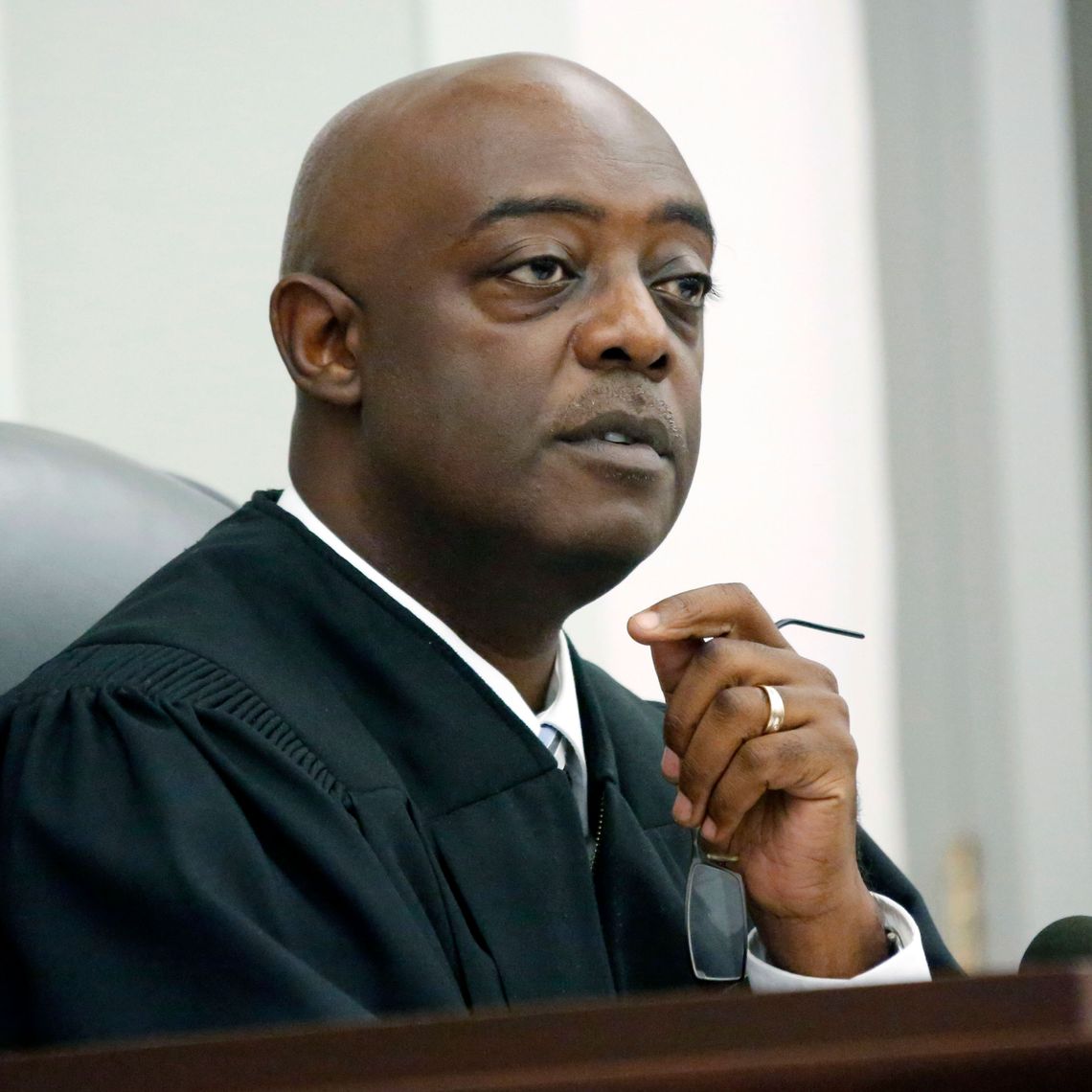 A bald, Black man wearing a black robe listens while holding his eyeglasses in his hand. 