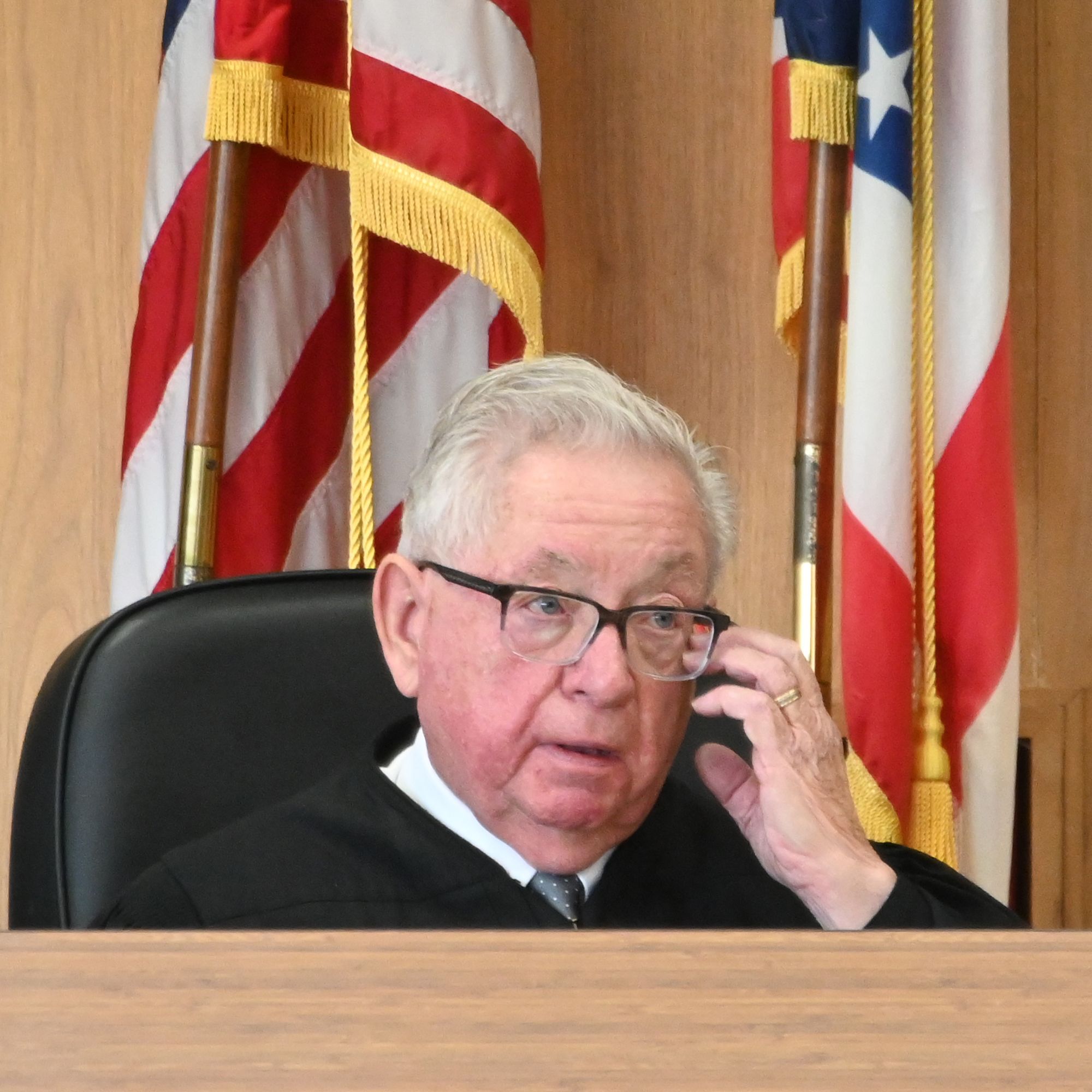Left: A photo shows Judge Mark Mihok, an elderly White man with glasses, with his hand raised to his face. He is wearing a judge’s robe, and American flags are standing behind him in the courtroom. 
