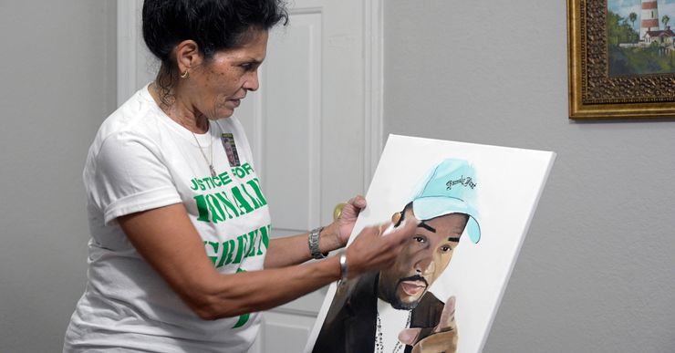 A woman with medium-toned skin and dark hair holds a painting of her son, Ronald Green, a Black man wearing a blue baseball cap.  