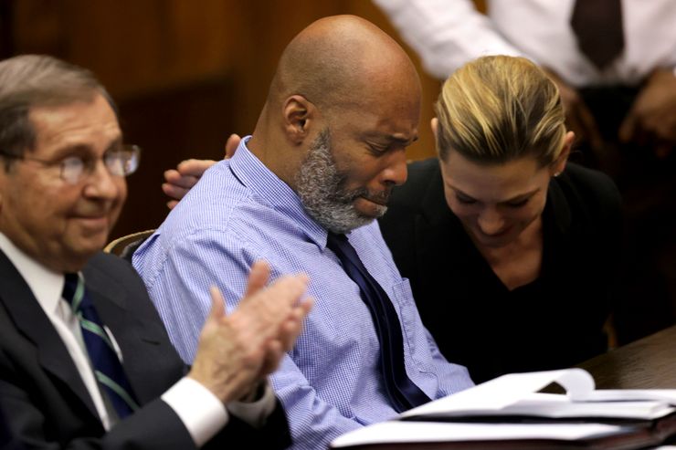 A Black man, wearing a blue striped button-down shirt and black tie, sits with an emotional look on his face and eyes closed. A White female lawyer, sitting on his right, is looking down and smiling while patting him on his back. A White male lawyer, sitting on his left, is smiling while clapping his hands. 