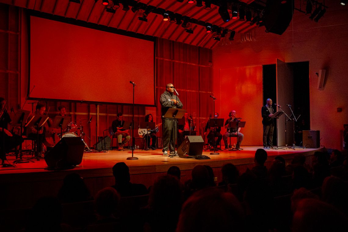A Black man with short, braided hair and sunglasses stands in the center of a stage wearing a black hooded sweatshirt and black pants. His hands are folded and rest on his chest. Behind him sits a row of seated musicians holding their instruments and a large movie screen on the wall. To the right of the stage, stands a bald Black man behind a microphone. Wearing black pants and a black suit coat over a grey shirt, he rests his hands on the edge a music stand in front of him.