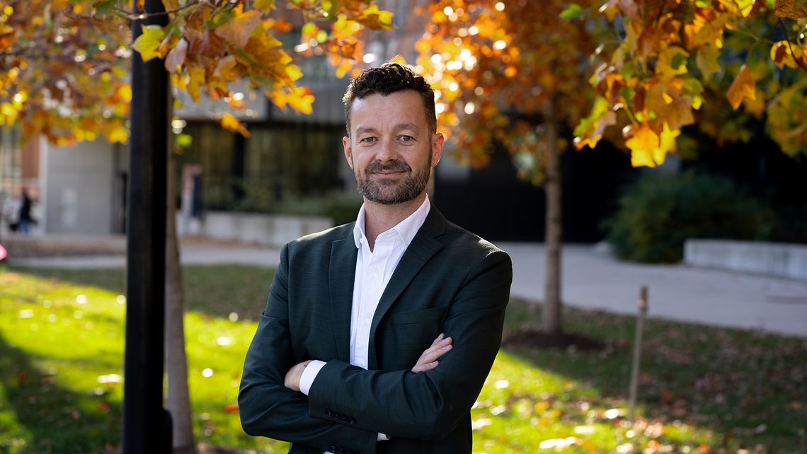 A White man in a white button-up shirt and black blazer stands with his arms crossed while posing for a portrait. 