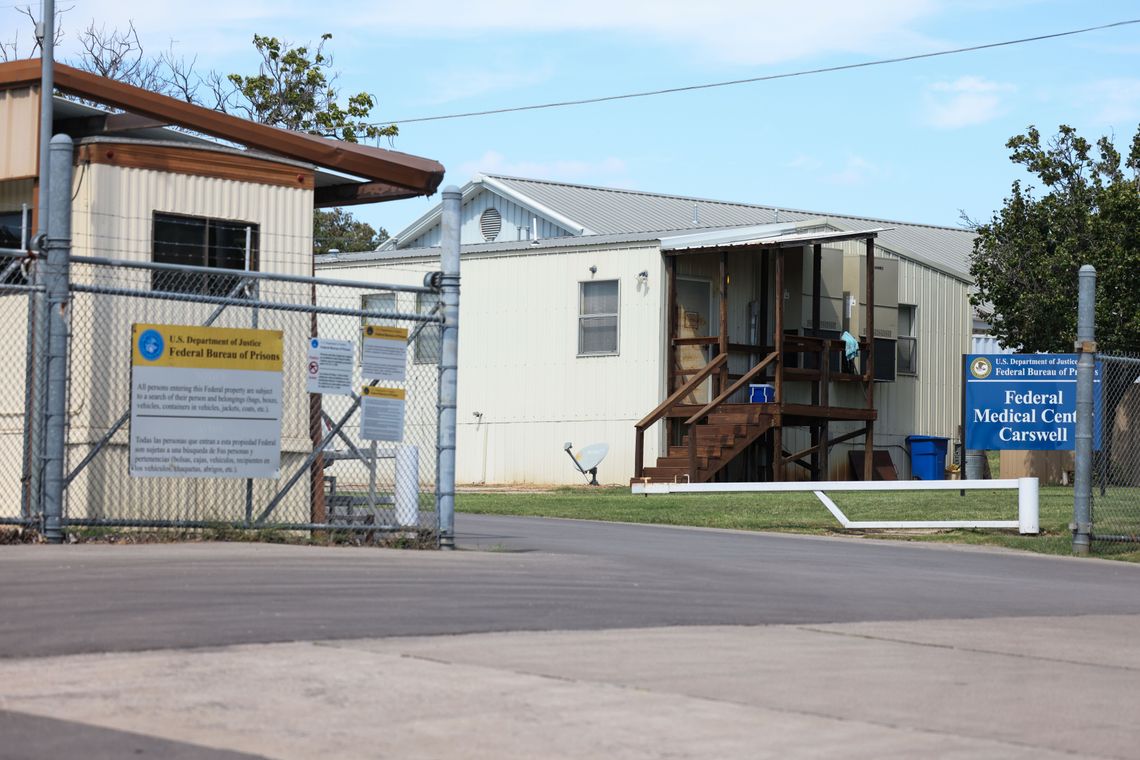 A photo shows metal fencing in front of two structures, one on the right with a short wooden staircase, and one on the left with beige siding. A sign in front of the building on the right reads: Federal Medical Center Carswell. 