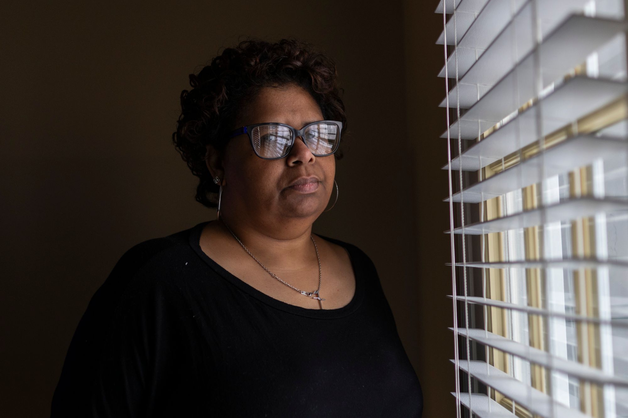 An older Black woman stands by a window looking at the camera. The blinds are reflected in her glasses.