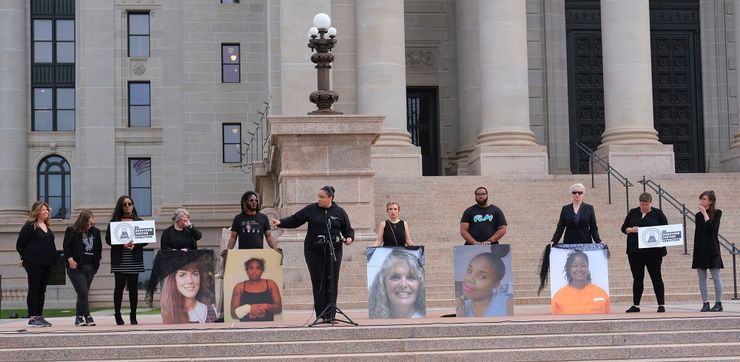 Demonstrators, dressed in black clothing, hold large posters of women and signs that read "OK Survivor Justice Coalition" while standing outside the Oklahoma State Capitol. 