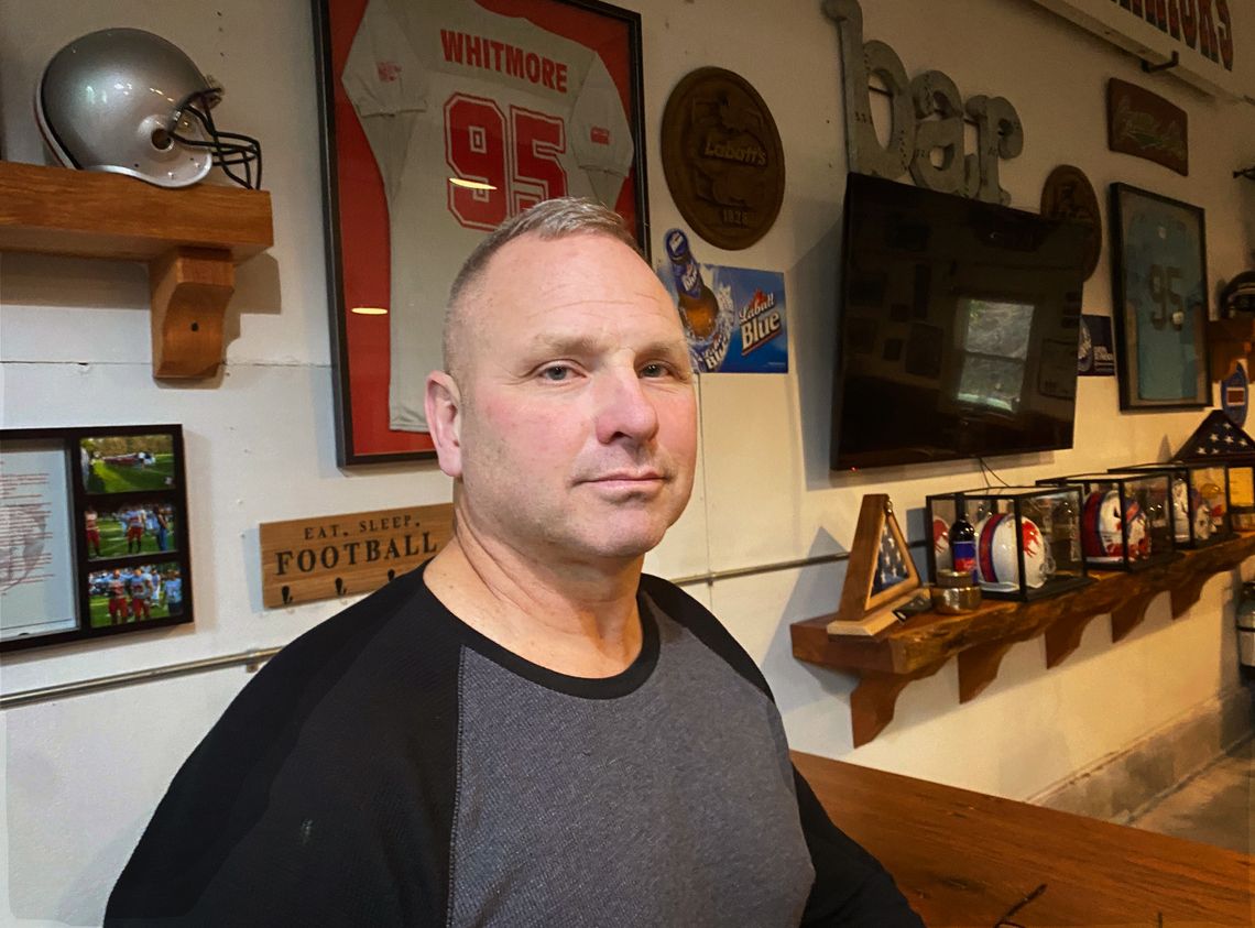 A White man with a neutral expression stands in front of a wall with sports memorabilia. 