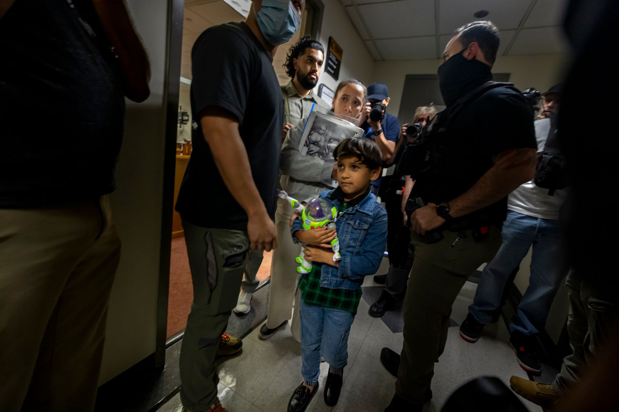 A photo shows a young boy with medium skin tone wearing a denim jacket and holding a Buzz Lightyear toy. A woman with medium-light skin tone stands behind him, and masked agents stand around them in a hallway.