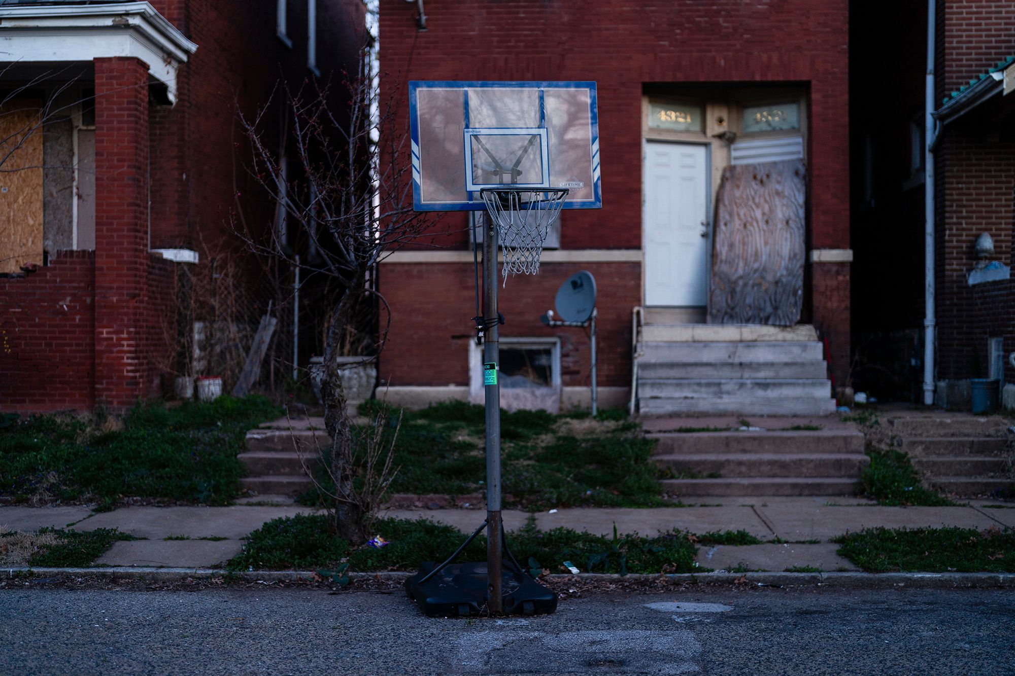 A photo shows a portable basketball hoop set up in a street, in front of a red house. 