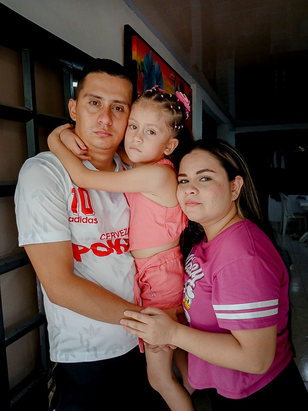 A photo shows a man with medium-toned skin and short dark brown hair holding a girl with medium-toned skin and dark brown hair, while his wife, a woman with medium-toned skin and dark brown hair, has her arm around them. 