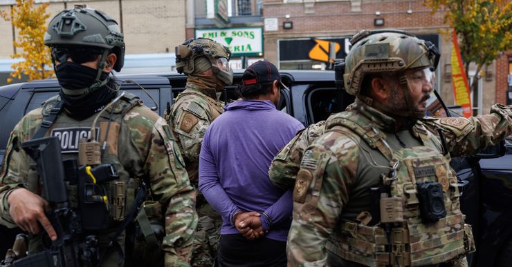 A man in a purple shirt has his hands cuffed behind his back while facing an open car door. Four federal officers, wearing camouflage uniforms, vests, and helmets, stand around the man. 