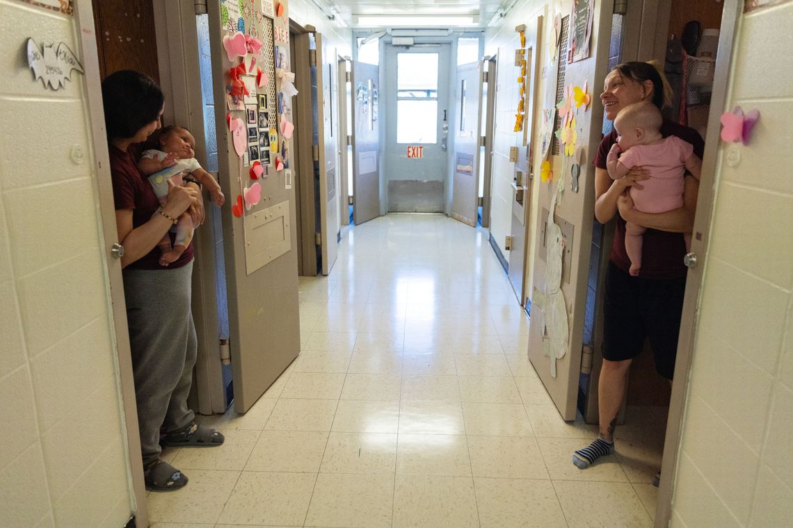 A White woman holding a baby stands in a doorway while looking toward another woman with a baby across the hallway. 