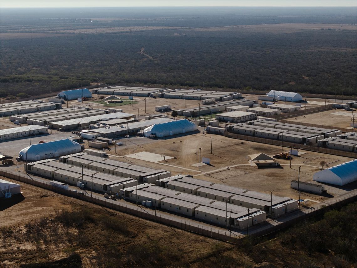 A photo shows an aerial view of a facility consisting of more than a dozen long rectangular buildings. The area around the facility has trees and brush. 