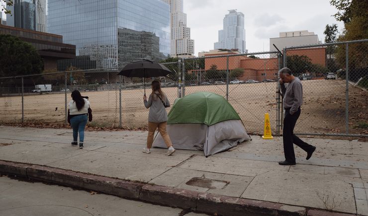 A photo shows Jazmine Mapes, a Native American woman with long dark hair, wearing a white sweater and jeans, walking near a chain link fence. An empty lot is situated behind the fence. In front of the fence is a tent. A woman holding an umbrella walks behind Mapes, and a man talking on a cell phone walks behind her.