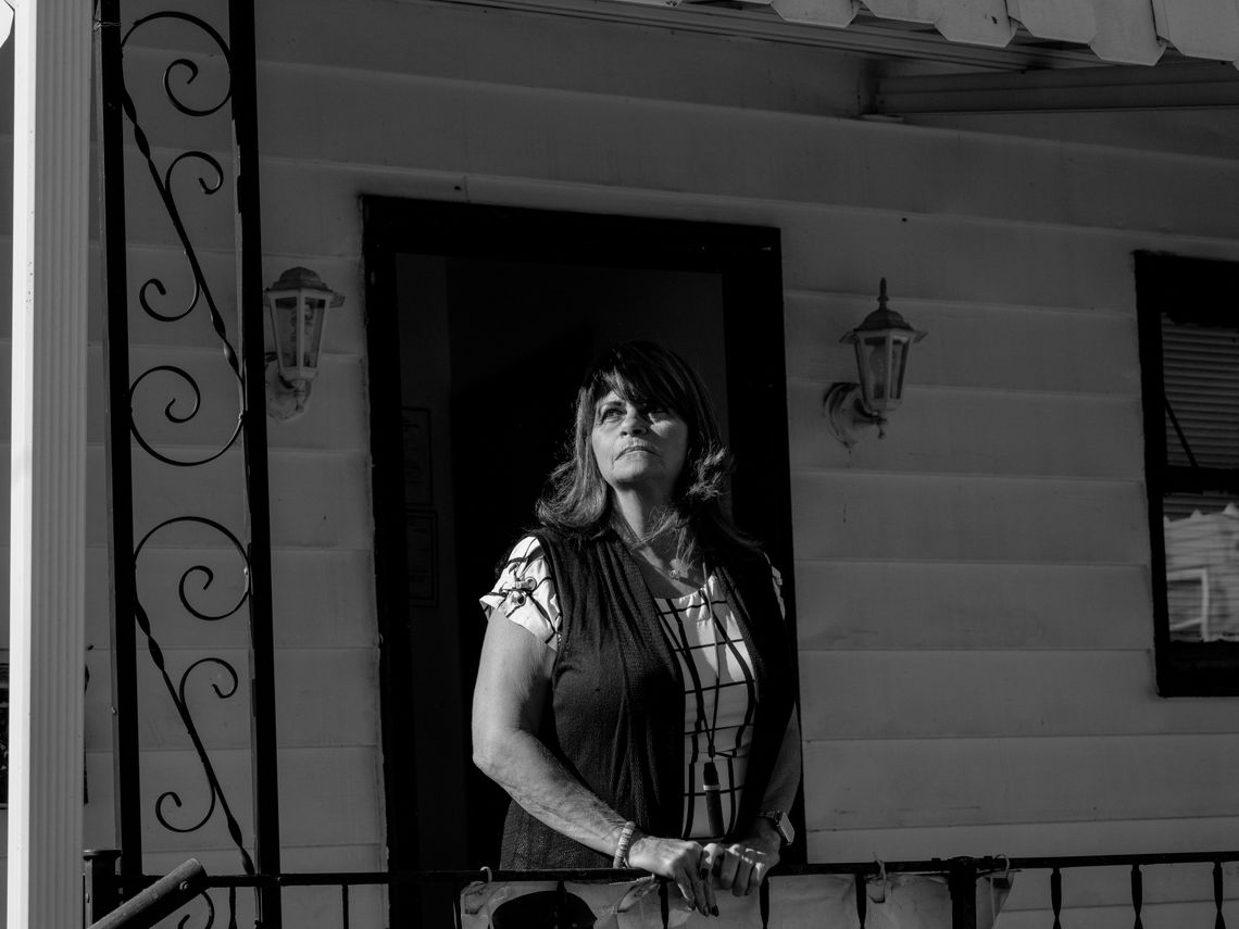 A black-and-white photo shows a woman with medium-length hair, a vest, and a short-sleeved shirt, standing in front of a building with white siding. 