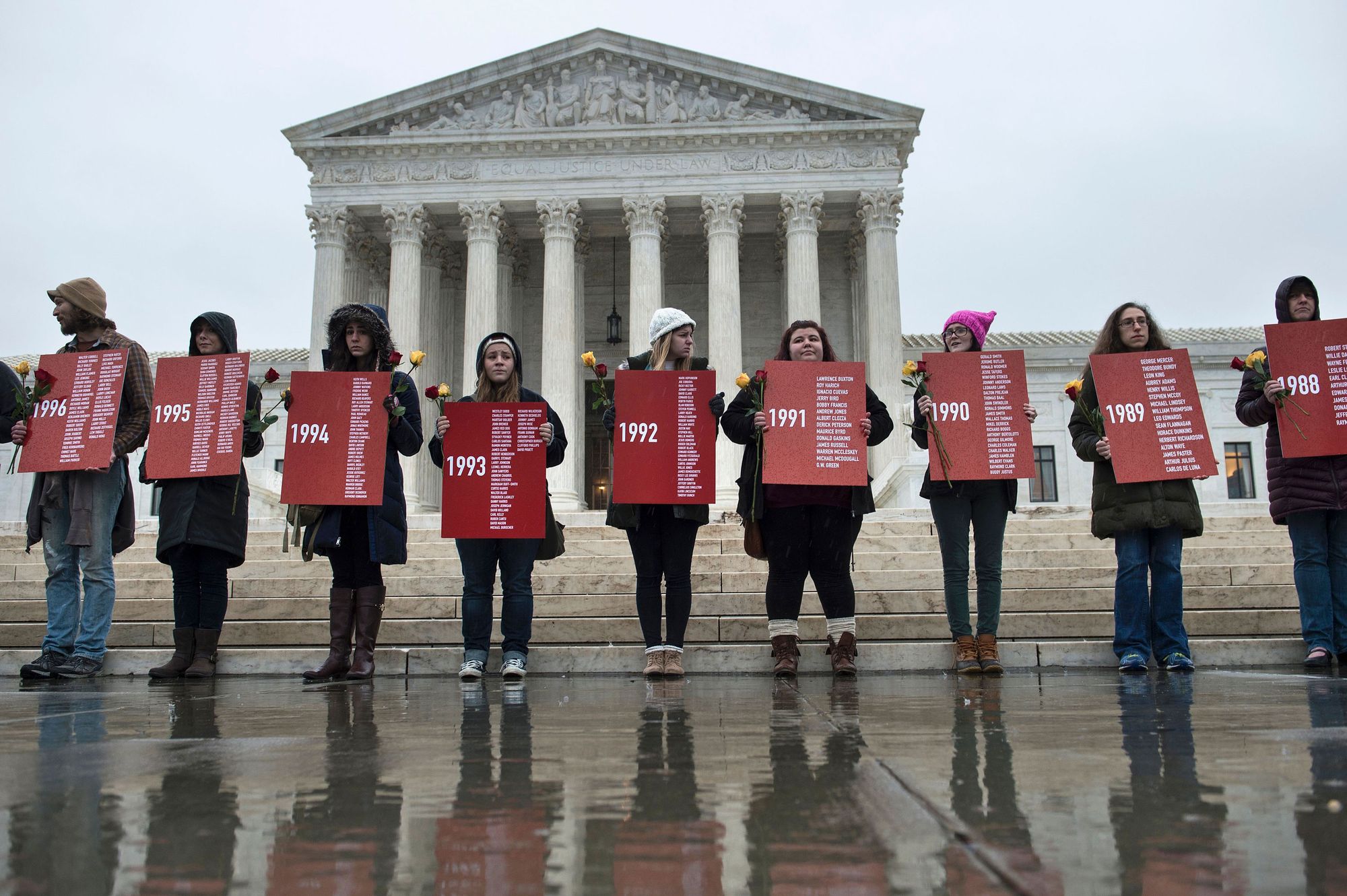 A photo of nine people holding red signs with dates and names in front of a white, columned marble building. 