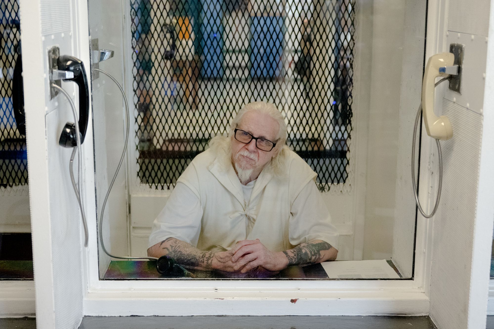 A photo shows a White man with long white hair and a beard, sitting behind glass in a booth in prison where visitors can speak to an incarcerated person. There is a phone on each side of the booth. Wood is wearing a white prison uniform. 